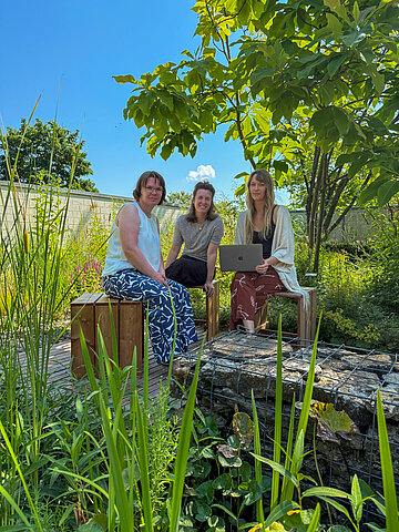 Photo: Three women are sitting in the shade of a tree by the pond on the campus in Höxter.