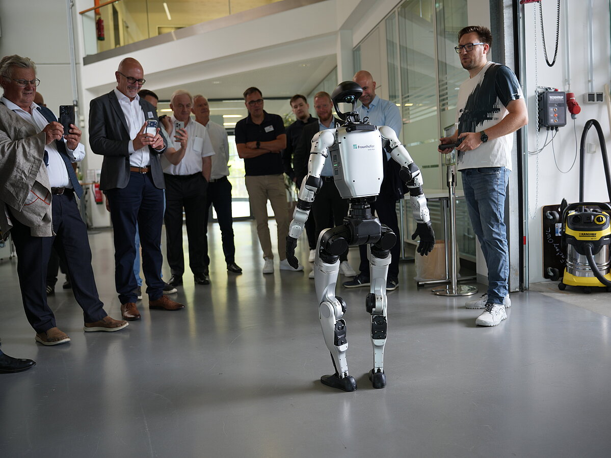 Der humanoide Roboter Karl steht in der Mitte einer Gruppe. Mehrere Mitglieder der Delegation richten ihre Smartphones auf ihn und fotografieren ihn.