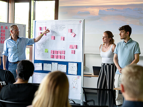 Photo: During a presentation, a man is standing to the left of a flip chart, while a woman and a man are standing on the right side of the picture.
