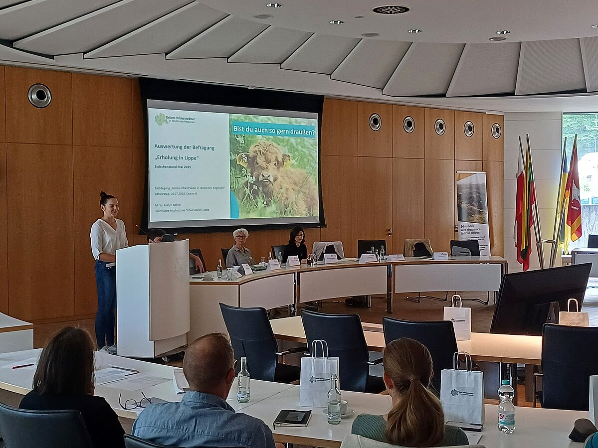 Tagungsteilnehmer:innen in einem Tagungsraum. Eine Dame hält einen Vortrag.