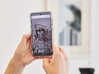 Zwei Hände halten ein Smartphone, auf dem Display ist ein Foto eines Kunstwerks zu sehen.