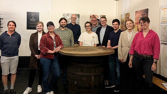 Eine Gruppe von etwa 10 Menschen aus dem Projekt KliMalz steht vor einem runden Tisch in der Brauerei Fiege und lächelt in die Kamera