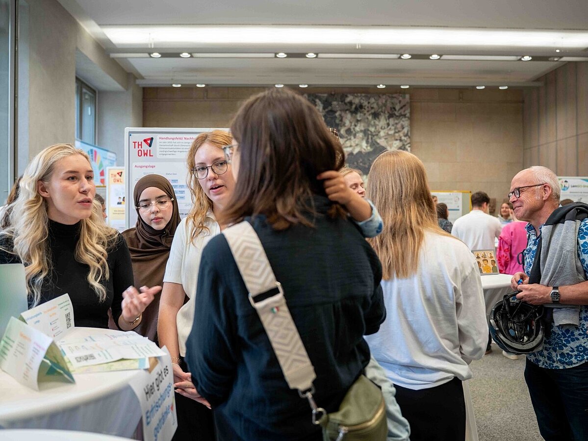 Mehrere junge Frauen stehen an einem Infostand und sprechen mit Besucherinnen und Besuchern. Auf dem Tisch liegen Flyer und Getränke. Im Hintergrund sind weitere Menschen und Poster mit Informationen der TH OWL zu sehen.