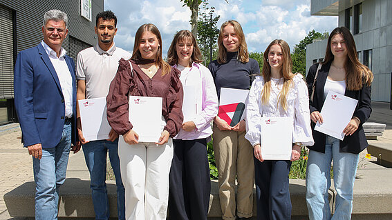 Seven people are standing together outside, looking toward the camera. Six of them are holding a document in their hands.