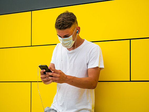 Junger Mann in weißem Shirt vor gelber Wand mit Mundschutz und Handy in der Hand
