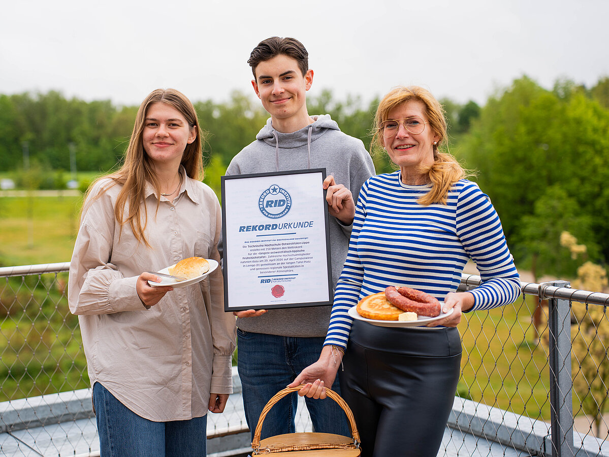 Zwei Frauen und ein Mann stehen zusammen. Der Hann steht in der Mitte und hält eine Urkunde in der Hand. Beide Frauen halten Teller mit Frühstück in der Hand. Die rechte Frau hält auch einen Frühstückskorb.