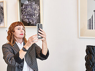 Prof.'in Stache hält ein Smartphone hoch und schaut aufs Display.