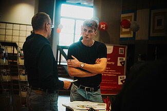 Junger Mann unterhält sich mit anderem Mann am Messestand.