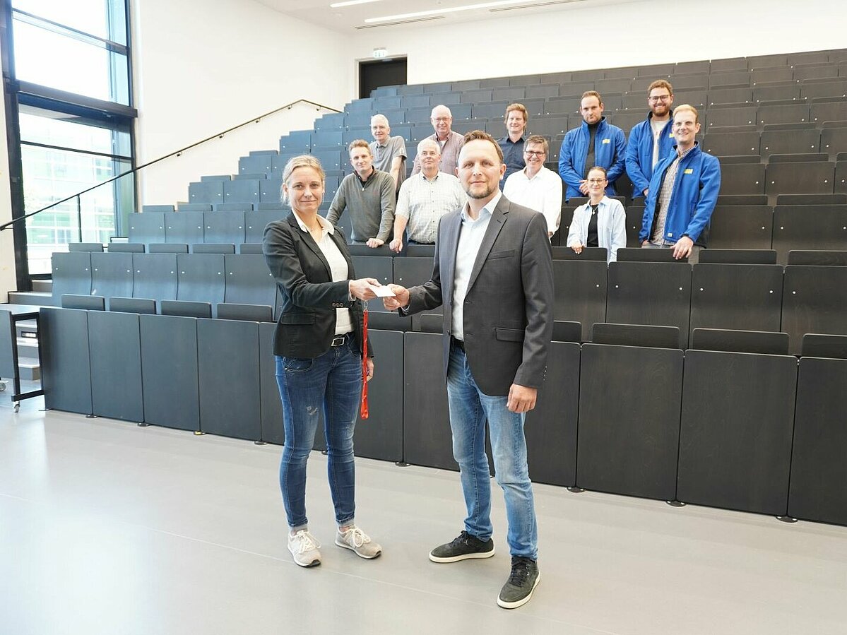 Hörsaal. Frau übergibt Schlüsselkarte an Mann. Einige Personen im Hintergrund.