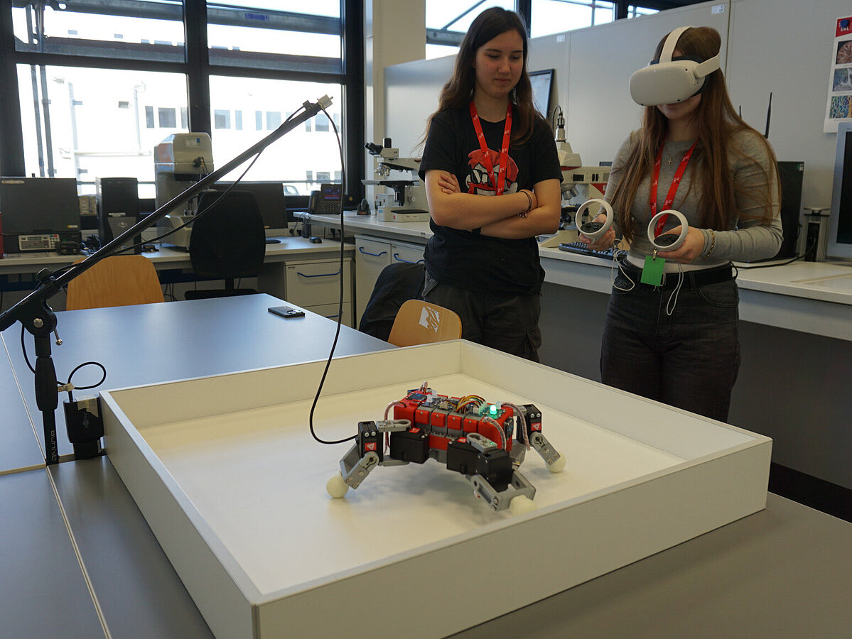 Ein Mädchen hat eine VR-Brille an und steuert einen Roboter. Ein weiteres Mädchen schaut ihr dabei zu. 