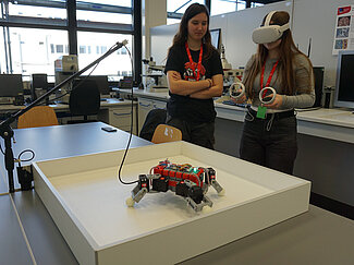 Ein Mädchen hat eine VR-Brille an und steuert einen Roboter. Ein weiteres Mädchen schaut ihr dabei zu. 