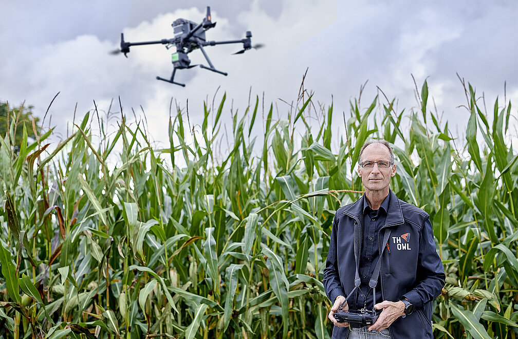 Eine landwirtschaftliche Drohne fliegt über ein Maisfeld. Prof. Dr. Wrenger steht davor mit Steuerungsgerät in den Händen.