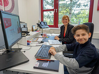 Ein Junge im dunklen Pullover sitzt im Vordergrund an einem Computer, Hände an Tastatur. Eine Frau sitzt im Hindergrund an demselben Schreibtisch.