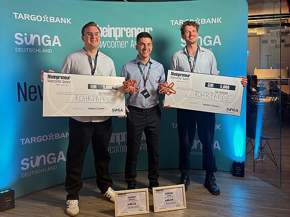 Drei junge Männer stehen lächelnd vor einer blau beleuchteten Wand mit Logos von Targobank, SINGA Deutschland und dem Rheinpreneur Newcomer Award. Zwei von ihnen halten übergroße Schecks über jeweils 5.000 Euro mit dem Namen ‚Rohrpappe‘. Alle tragen Businesskleidung und haben Medaillen oder Namensschilder um den Hals. Vor ihnen auf dem Boden stehen gerahmte Urkunden. Der Raum hat eine moderne Atmosphäre mit sichtbaren Lüftungsrohren an der Decke.