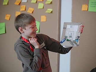 Ein Junge hält ein Stück Pappe hoch, auf der er aus vielen verschiedenen Zeitungsschnipseln einen Raum gestaltet hat.