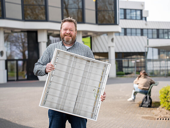 Ein Mann präsentiert ein ausgebautes Leuchtpanel vor dem Hauptgebäude des Campus.
