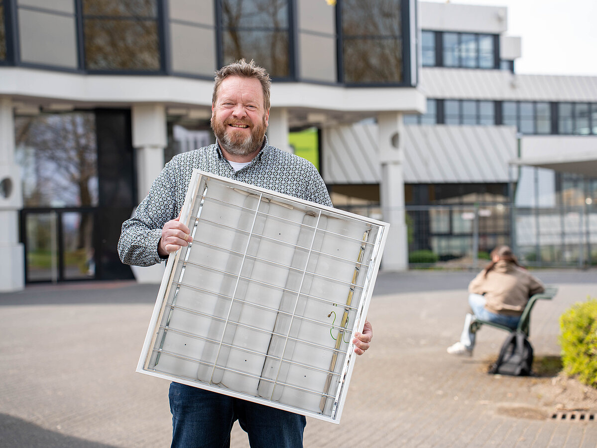 Ein Mann präsentiert ein ausgebautes Leuchtpanel vor dem Hauptgebäude des Campus.