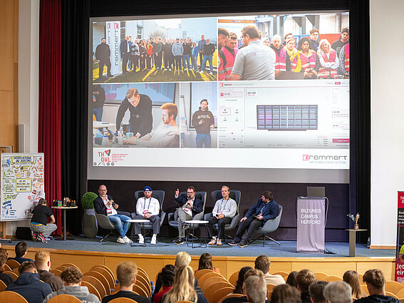 Blick aus dem Publikum auf eine Podiumsdiskussion auf der DiMS-Konferenz am BildungsCampus Herford. Auf der Bühne sitzen mehrere Gesprächsteilnehmende im Austausch, während auf einer Leinwand Bilder eines Ideationsprints gezeigt werden. Am rechten Bildrand erstellt eine Graphic Recorderin eine visuelle Zusammenfassung der Veranstaltung.