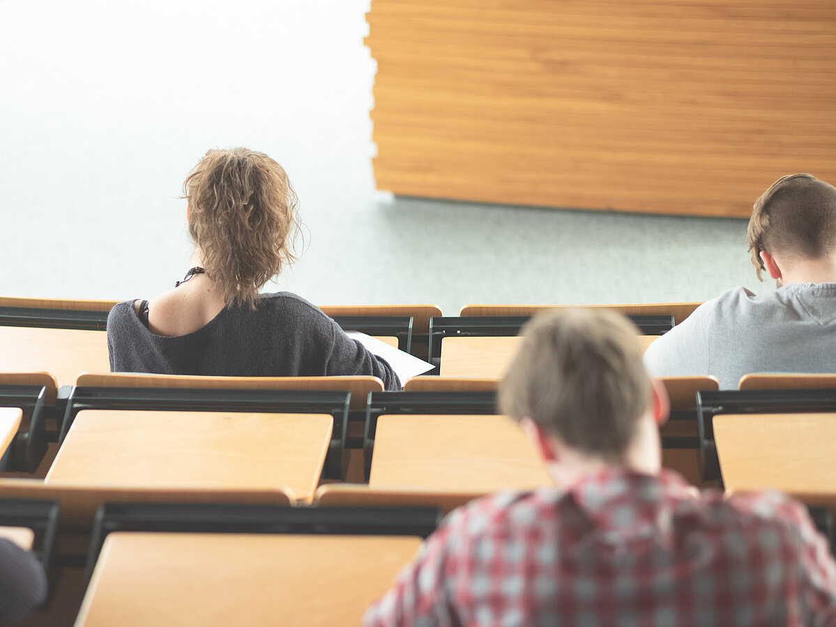 Mehrere Studierende sitzen in einem Hörsaal und arbeiten an ihren Unterlagen; die Aufnahme zeigt sie von hinten in einer ruhigen Lernsituation.