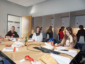 Drei Mädchen sitzen an einem Tisch und arbeiten. Vier andere sitzen in Kabinen, um einen Geruchs- beziehungsweise einen Geschmackstest durchzuführen.