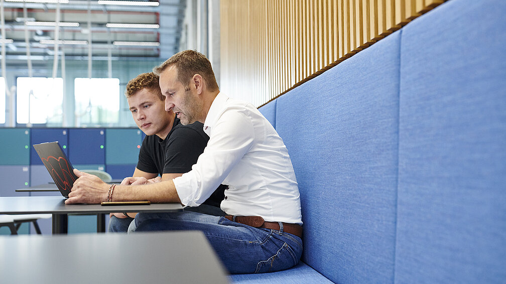 Prof. Dr. Paa sitzt mit einem jungen Mann an einem Cafeteriatisch. Beide schauen auf das Display eines Laptops.