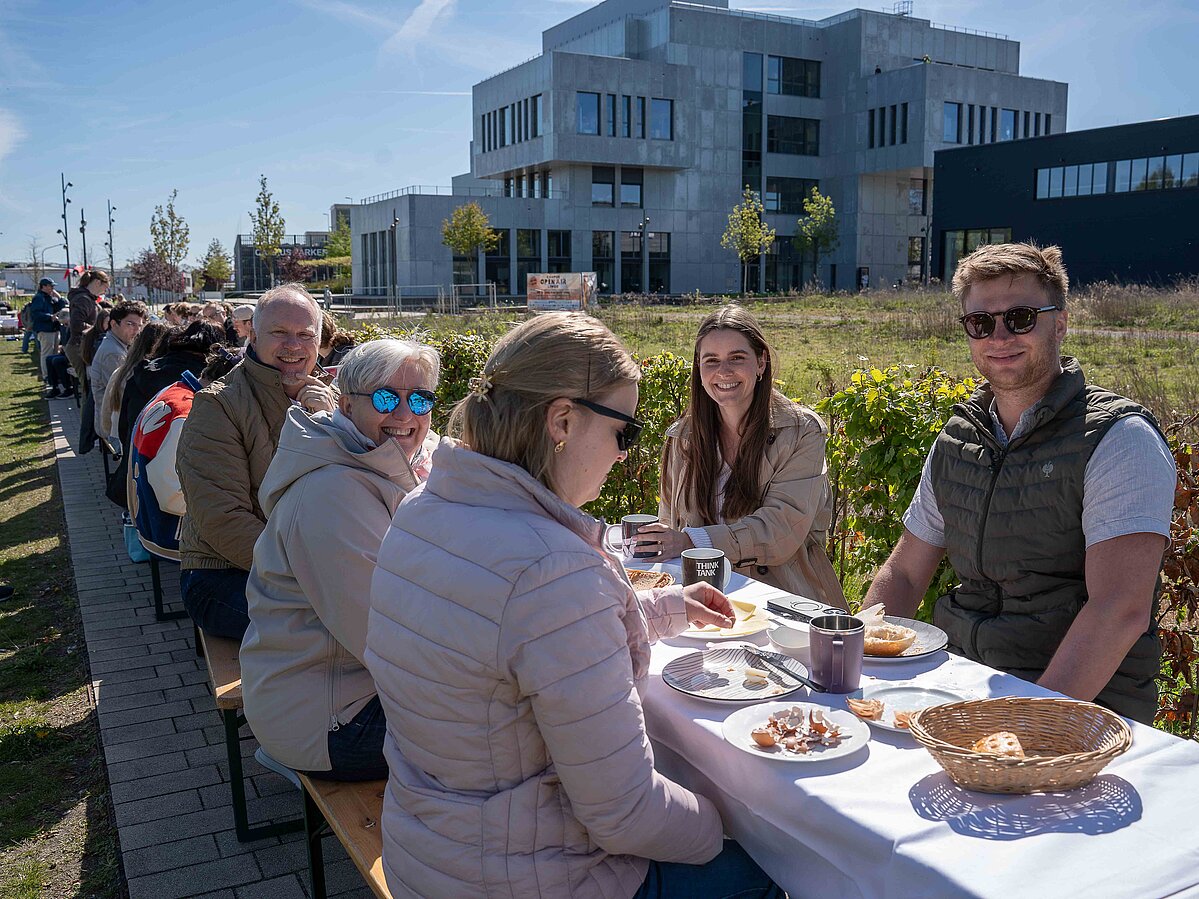Mehrere Menschen sitzen bei sonnigem Wetter an einer langen gedeckten Frühstückstafel auf dem Campus und essen gemeinsam Brötchen und trinken Kaffee vor einem modernen Hochschulgebäude.