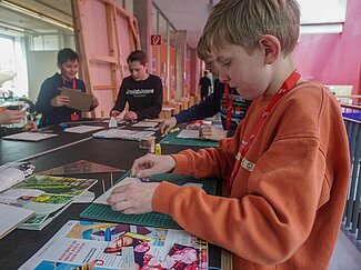 Ein Junge im organgenen Sweatshirt im Vordergrund und drei weitere Jungs im Hintergrund erstellen eine Kollage aus Zeitungsausschnitten.