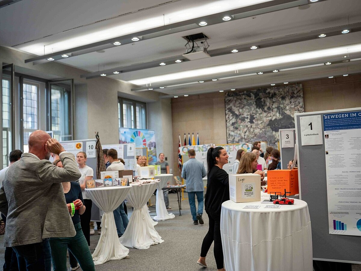 Menschen besuchen eine Ausstellung in einem großen Saal. An mehreren Stehtischen und Stellwänden präsentieren Studierende der TH OWL verschiedene Projekte und Poster. Besucherinnen und Besucher schauen sich die Infotafeln an, tauschen sich aus oder diskutieren in kleinen Gruppen.
