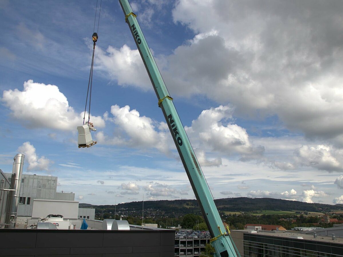Ein Kranarm hat eine Mischmaschine am Haken und will diese auf dem Dach absetzen.