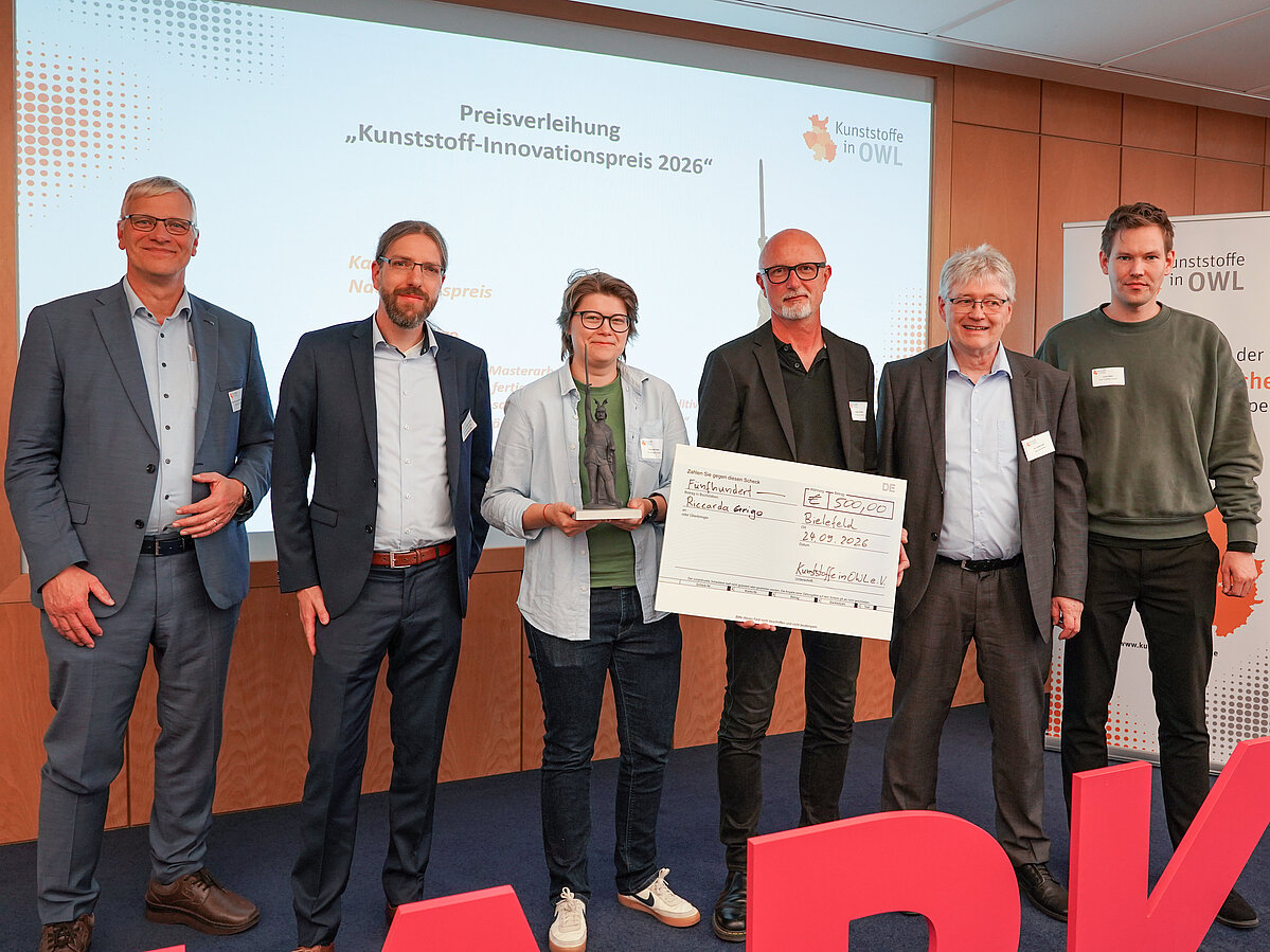 Photo: Five men and one woman are standing side by side, looking at the camera; a presentation slide is visible in the background. One of the men in the group is holding up a symbolic check. 