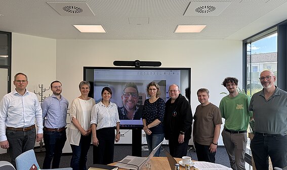 UrbanMobilPlus: Adaptive Mobilitätsplattform für Stadt, Quartier und Campus Foto: Zehn Personen, davon drei Damen, sind auf in einem Seminarrraum in einer Reihe nebeneinander zu sehen. Ein Mann ist auf dem großen Monitor dahinter zugeschaltet.