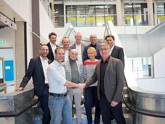 Foto: Eine Gruppe von Menschen steht auf einer Treppe. Ganz vorn stehen zwei Männer, die sich die Hände schütteln und lächeln.