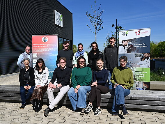 A group of participants is standing and sitting in front of the Future Food Factory OWL on the Innovation Campus Lemgo. Next to them are roll-up banners showcasing the SIXFOLD and Future Food Factory OWL projects. The group is posing for a group photo in sunny weather as part of a study visit focused on digital transformation in the food industry.