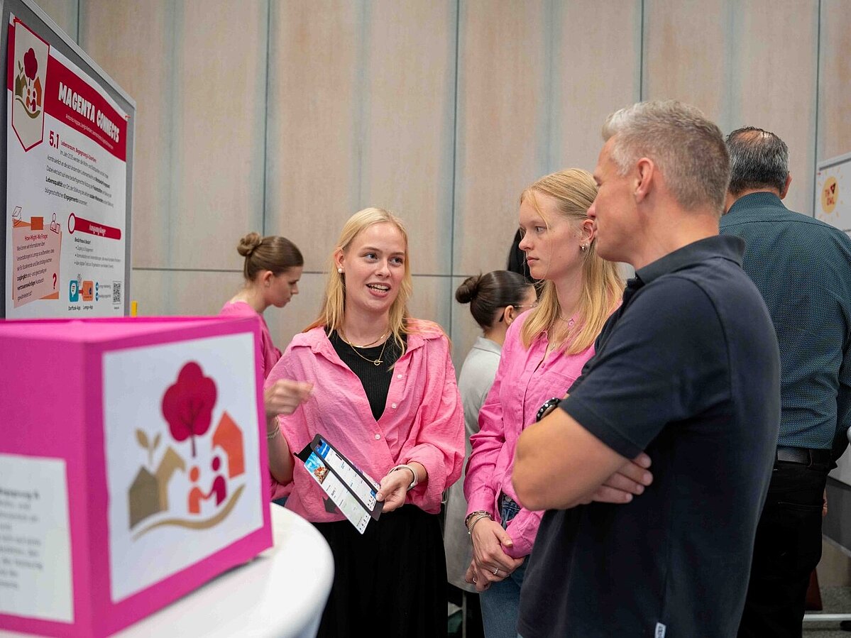 Zwei junge Frauen in pinken Hemden sprechen an einem Infostand mit einem Mann. Eine der Frauen hält einen Flyer in der Hand und erklärt etwas. Im Hintergrund sind Poster und Stellwände mit Projektinformationen sowie weitere Personen zu sehen.