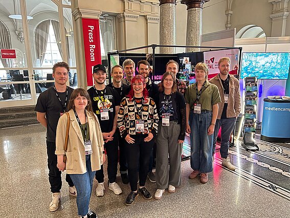 A group of students, staff, and professors from the Faculty of Media and Culture at TH OWL is standing in front of the university’s booth at FMX 2026 in Stuttgart. In the background, a red “Press Room” sign, monitors, informational materials, and exhibition stands are visible.