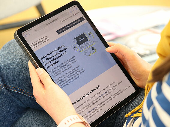 Blick über die Schulter einer Studentin auf ein Tablet mit der geöffneten Internetseite zur Energiepreispauschale.
