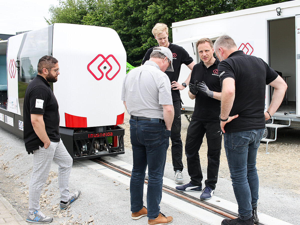 Das Forschungsteam steht auf der Strecke zusammen und bespricht die Testfahrt. Dahinter steht das MONOCAB.