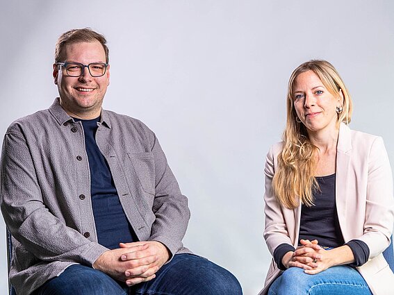 Zwei Personen, ein Mann und eine Frau sitzen vor einer weißen Wand auf Stühlen. 