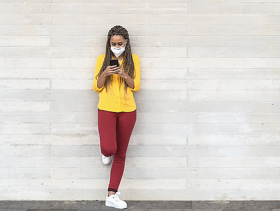 Junge Frau im gelben Shirt mit Mundschutz und Smartphone vor einer grauen Wand