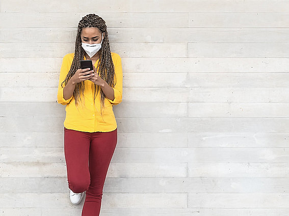 Frau mit Maske und Smartphone lehnt sich an eine Wand