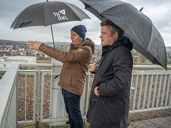 Foto: Zwei Männer mit ausfgespannten Regenschirmen blicken von der Dachterrasse des TH-OWL-Hauptgebäudes.