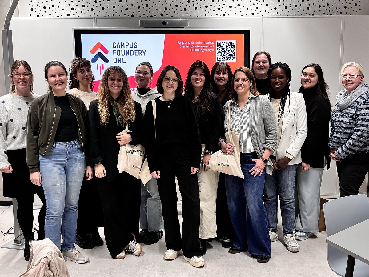 Photo: A group of 13 women are standing in a seminar room and looking at the camera. In the background, there is a large monitor on the wall. It shows the logo of Campus Foundery OWL.