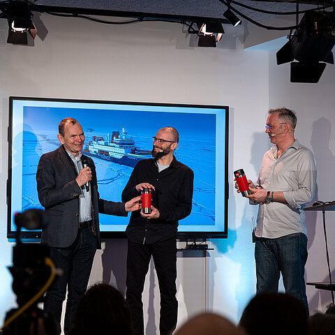 Photo: Three men standing on a stage. A display behind them shows an image of the research vessel Polarstern in the ice.