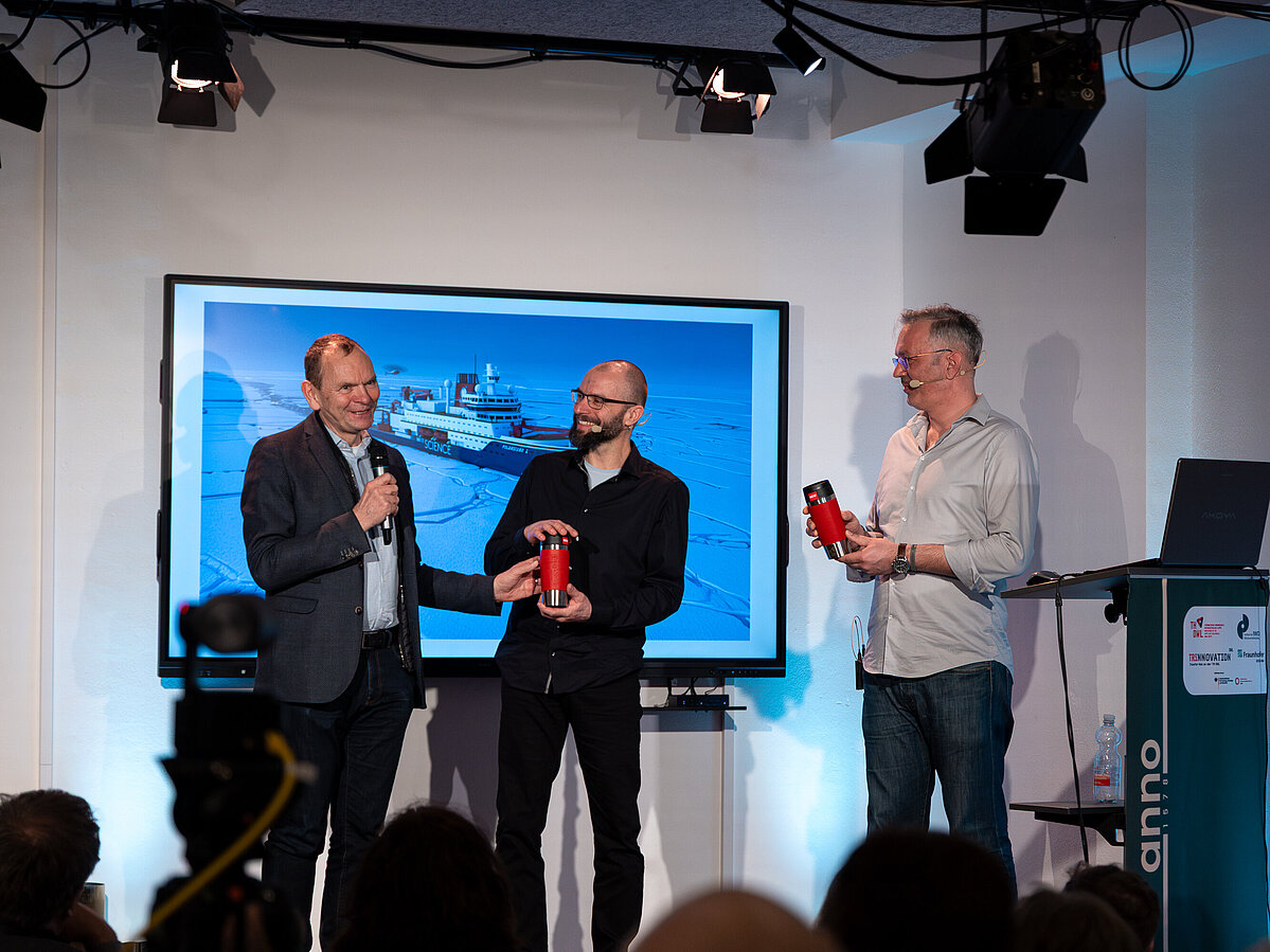 Forschen in „menschenfeindlichem Gebiet“ Foto: Drei Männer stehen auf einer Bühne. Ein Display hinter ihnen zeigt ein Bild des Forschungsschiffs Polarstern im Eis..