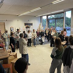 Ansammlung von jungen Menschen die im Kreis stehen in einem Raum, eine Person spricht in ein Mikrofon.