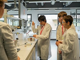 Drei Jungen im weißen Kittel im Chemielabor. Zwei Jungen schauen auf den dritten, der mit einer großen Pipette arbeitet.