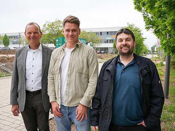 Three men are standing side by side outdoors at the Innovation Campus Lemgo, looking into the camera. In the background, you can see buildings, trees, and a construction site.