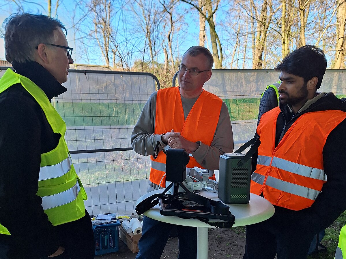 Drei Männer diskutierend um Stehtisch im Freien.