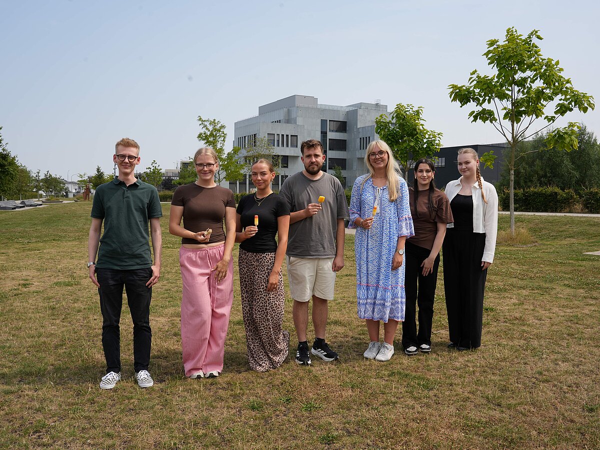 Foto: Eine Gruppe von sieben Personen, fünf Frauen und zwei Männer, steht auf dem Campuswiese in Lemgo. Im Hintergrund in einiger Entfernung steht das Gebäude InnovationSPIN.