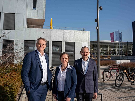 Drei Personen in Businesskleidung stehen bei sonnigem Wetter vor einem modernen Hochschulgebäude auf dem Campus der Technischen Hochschule Ostwestfalen-Lippe und posieren für ein Gruppenfoto. Fahrräder und weitere Campusgebäude sind im Hintergrund zu sehen.